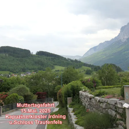 Eine malerische Route am 15. Mai 2025 führt zum Kapuzinerkloster Irdning und Schloss Trautenfels. Der Weg wird von üppigem Grün und einer Steinmauer gesäumt. Im Hintergrund sind Berge und ein Dorf zu sehen.