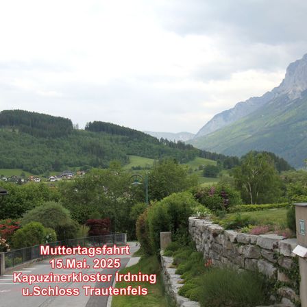 Eine malerische Route am 15. Mai 2025 führt zum Kapuzinerkloster Irdning und Schloss Trautenfels. Der Weg wird von üppigem Grün und einer Steinmauer gesäumt. Im Hintergrund sind Berge und ein Dorf zu sehen.