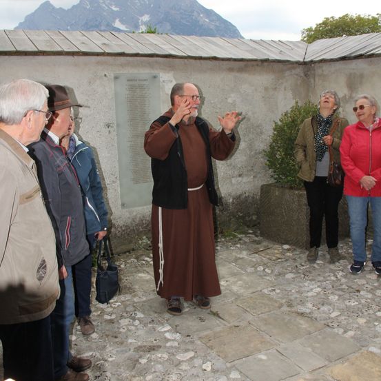 Ein Mann in einer braunen Robe spricht zu einer Gruppe von Menschen, die um eine Mauer mit einer Gedenktafel und Pflanzen stehen.