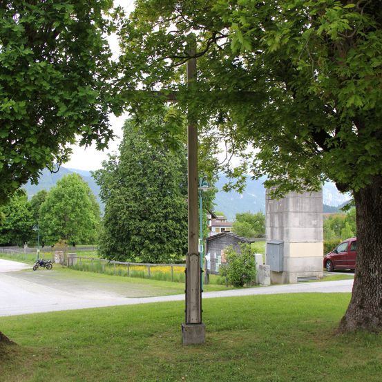 Ein kleines Straßenkreuz steht zwischen üppigen Bäumen, mit einem geparkten Motorrad, einem Gebäude und einem roten Auto im Hintergrund.