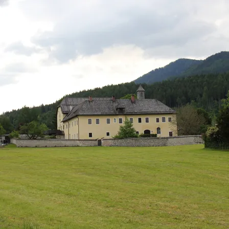 Ein großes gelbes Gebäude mit einem grauen Dach und mehreren Fenstern befindet sich in einem weitläufigen offenen Feld mit einer Steinmauer und üppiger Vegetation, vor der Kulisse einer bergigen Landschaft.