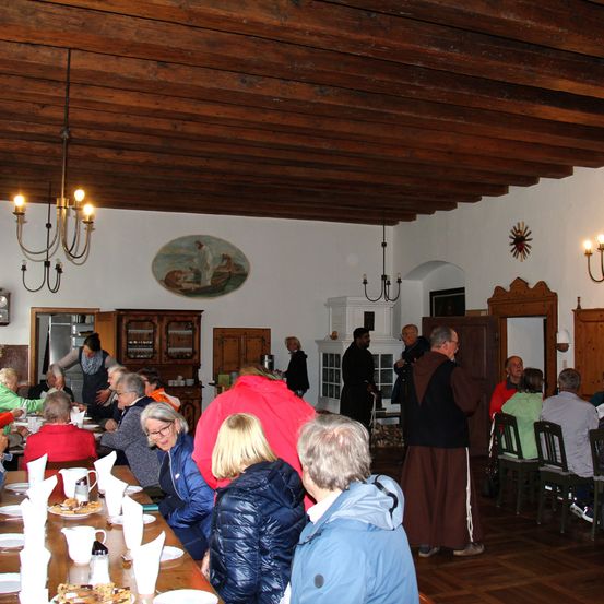 Ein großer Speisesaal mit Holzdecke und Wänden. Im Vordergrund befindet sich ein Tisch mit Tellern, Tassen und Essen. Menschen sitzen und stehen um den Tisch herum.