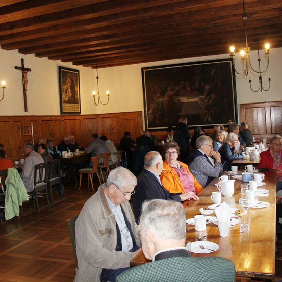 Ein Innenspeisesaal mit Holzwänden und einem Holzboden. Es gibt Tische mit Stühlen, Tassen, Tellern und Servietten. Ein großes Gemälde an der Wand. Eine Kronleuchter hängt von der Decke.