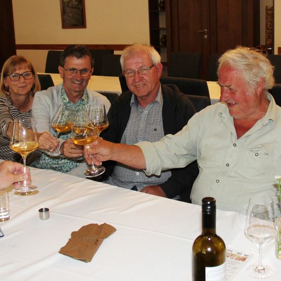 Eine Gruppe von vier Personen stößt mit Weingläsern an einem Tisch an. Ein Mann trägt eine Brille und eine graue Jacke. Hinter ihnen sind Stühle um einen Tisch herum angeordnet.