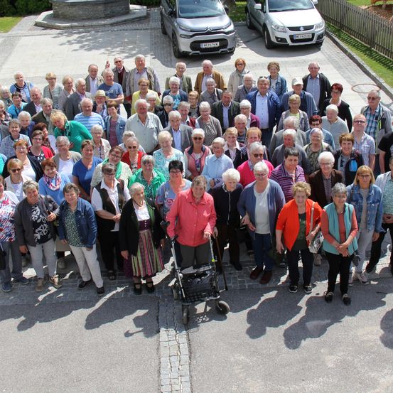 Eine große Gruppe von Menschen ist im Freien versammelt, viele tragen Sonnenbrillen. Sie stehen auf einem gepflasterten Bereich mit geparkten Autos dahinter. Die Atmosphäre scheint sozial und festlich zu sein.