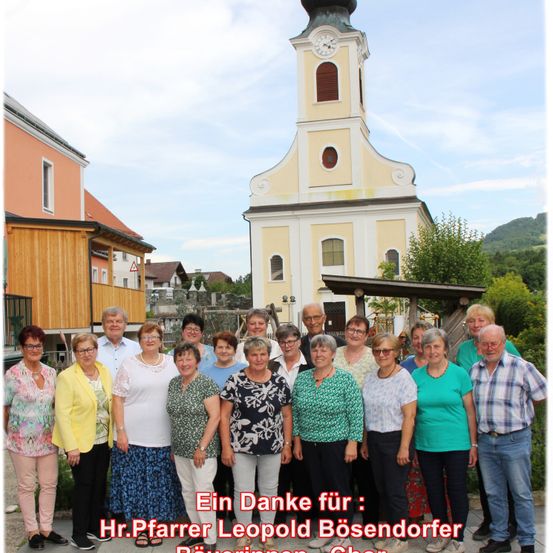 Eine Gruppe von Menschen posiert für ein Foto vor einer Kirche. Der Text lautet 'Ein Danke für:'. Die Kirche hat einen Turm mit einer Uhr. Bäume und Berge sind im Hintergrund zu sehen.