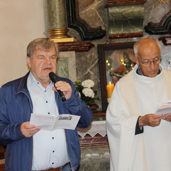 Ein älterer Mann spricht in ein Mikrofon und hält ein Buch in einer Kirche mit einer Kerze und religiöser Kunst.