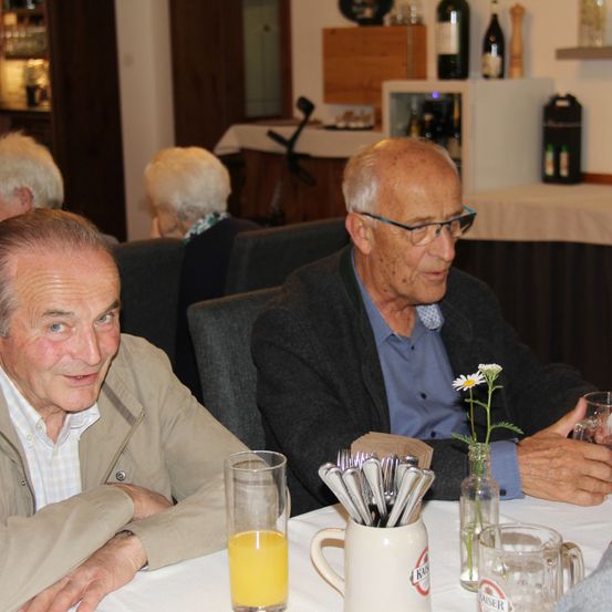 Zwei ältere Männer sitzen an einem Tisch in einem Restaurant, einer hält ein Glas Orangensaft und der andere ein Glas Wein. Hinter ihnen sitzen andere Gäste an Tischen.