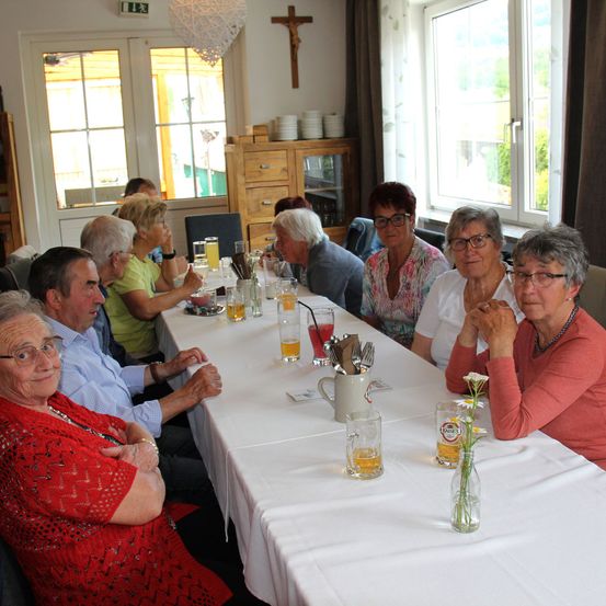Eine Gruppe älterer Menschen sitzt an einem langen Tisch in einem Essbereich. Sie trinken Bier und es gibt Blumen in einer Vase. Hinter ihnen sind Fenster mit Vorhängen.