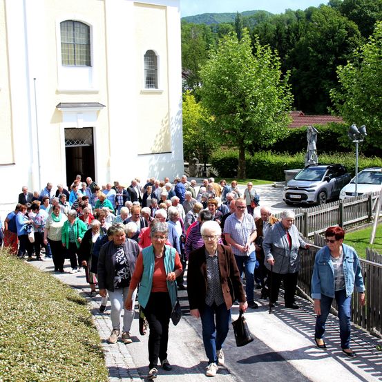 Eine Gruppe älterer Menschen geht auf einem Weg vor einer Kirche, mit einem geparkten Auto in der Nähe.