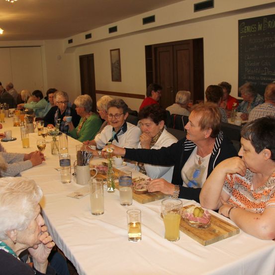 Eine Gruppe älterer Frauen sitzt um einen langen Tisch und unterhält sich. Sie genießen eine Mahlzeit, mit verschiedenen Getränken und Essensartikeln auf dem Tisch.