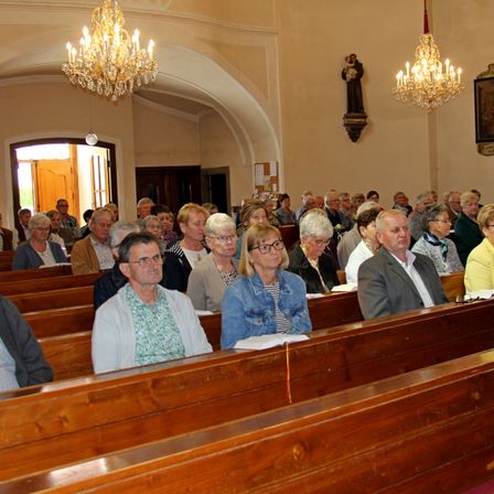 Eine Gemeinde älterer Menschen sitzt aufmerksam in einer Kirche. Sie sind in formeller Kleidung gekleidet und halten Bücher. Der Raum ist mit Holzbänken, Kronleuchtern und einer Statue eines Heiligen geschmückt.