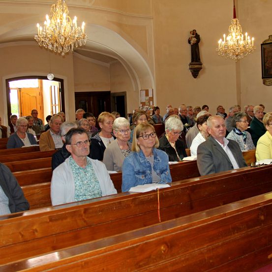 Eine Gemeinde älterer Menschen sitzt aufmerksam in einer Kirche. Sie sind in formeller Kleidung gekleidet und halten Bücher. Der Raum ist mit Holzbänken, Kronleuchtern und einer Statue eines Heiligen geschmückt.