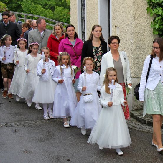Eine Gruppe von Kindern in weißen Kleidern geht mit Erwachsenen auf einem Weg, hält Kerzen und Blumen. Ihnen folgen mehrere Erwachsene, einige mit Brille. Bäume und ein Gebäude sind im Hintergrund.