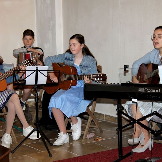 Vier junge Musiker sitzen auf Stühlen in einem Raum. Drei von ihnen spielen Gitarre, einer spielt Akkordeon. Der Keyboarder sitzt auf einem Schemel und spielt ein Roland-Keyboard.