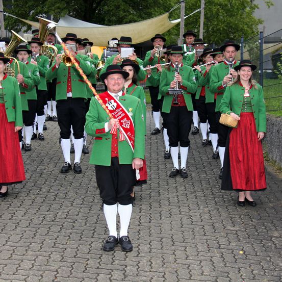 Eine Blaskapelle in traditionellen grünen und roten Uniformen spielt Instrumente auf einem gepflasterten Weg. Ein Mann mit einem Dirigentenstab führt die Band an.