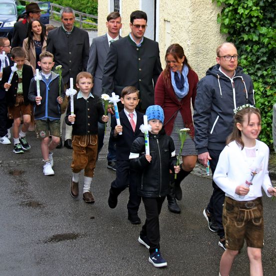 Eine Gruppe von Kindern und Erwachsenen geht einen Pfad hinunter, einige halten Blumen. Sie scheinen in einer Prozession zu sein, mit gemischter formeller und lässiger Kleidung.