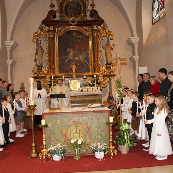 Ein Priester leitet einen Gottesdienst an einem Kirchenaltar, vor ihm halten Kinder Kerzen. Hinter dem Altar befindet sich ein großes Gemälde.