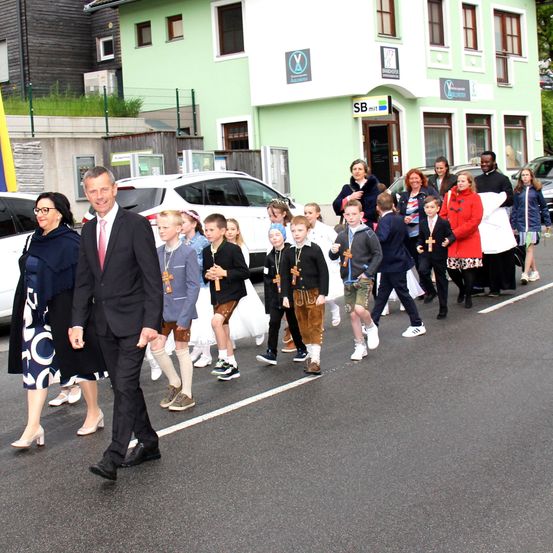Eine Gruppe von Menschen, darunter Kinder in traditioneller Kleidung, geht mit einem Mann in Anzug an der Spitze die Straße entlang. Dahinter ist ein grünes Gebäude mit Beschilderung zu sehen, und mehrere Autos sind geparkt.