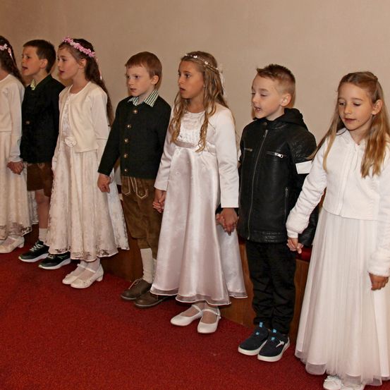 Eine Gruppe von Kindern, einige in weißen Kleidern und andere in Freizeitkleidung, stehen auf einem roten Teppich und nehmen wahrscheinlich an einer Schulveranstaltung teil.