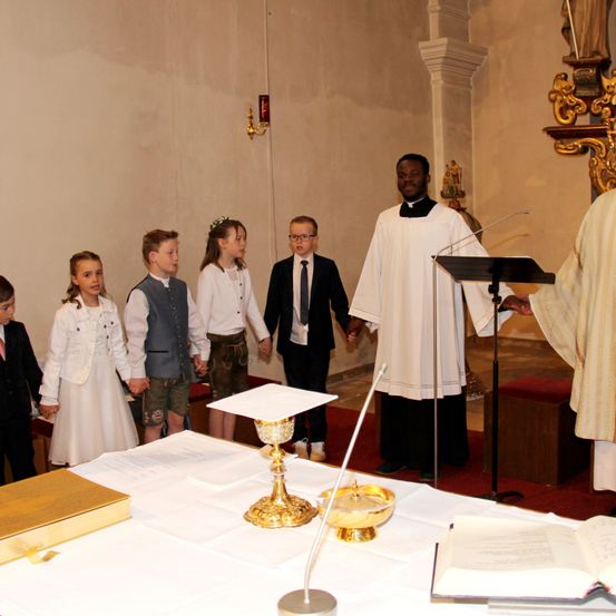 Mehrere Kinder in formeller Kleidung halten während einer Zeremonie in einer Kirche die Hände mit zwei Priestern. Ein Tisch mit einem Buch und einem Kelch steht vor ihnen.