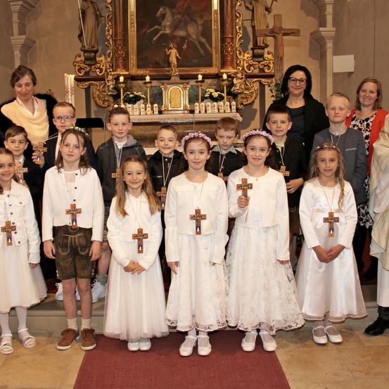 Eine Gruppe von Kindern in weißen Kleidern mit Kreuzen auf ihren Kleidern steht in einer Kirche. Sie posieren für ein Foto mit Erwachsenen hinter ihnen.