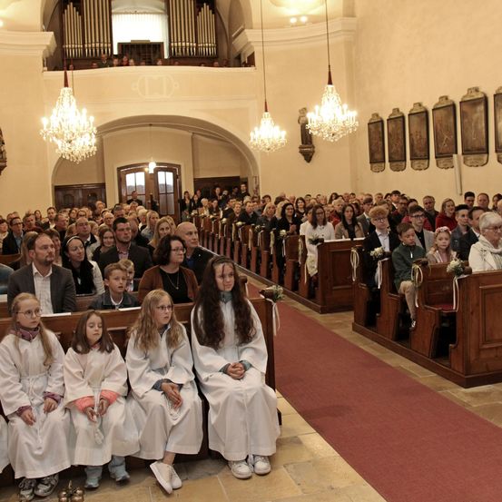 In einer Kirche versammelt sich eine große Gruppe von Menschen. Vier junge Mädchen in weißen Roben sitzen in der ersten Reihe. Die Kirche ist mit Kronleuchtern und Gemälden geschmückt.