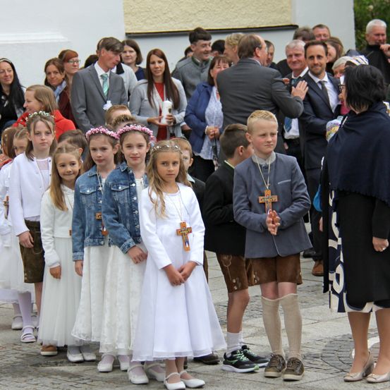 Eine Gruppe von Kindern in weißen Kleidern, einige mit Blumen im Haar, stehen in einer Reihe, wahrscheinlich für eine religiöse Zeremonie. Sie tragen Halsketten mit Kreuzen. Dahinter stehen Erwachsene in formeller Kleidung, einige klatschen.