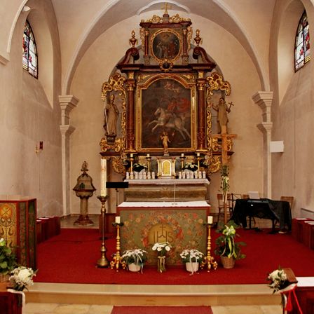 Ein kunstvoller Altar mit einer religiösen Malerei, Kerzen und Blumenarrangements schmückt das Innere einer Kapelle. Der Altar hat ein aufwendiges Design mit Statuen und goldenen Akzenten.