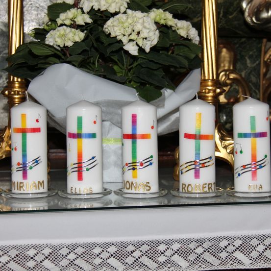 Fünf weiße Kerzen mit bunten Musiknoten und Namen wie Miriam, Elias, Jonas, Robert und Eva stehen vor einem goldenen Rahmen. Hinter den Kerzen befindet sich ein weißer Blumenstrauß.