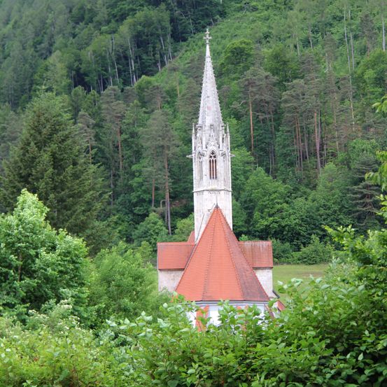 Eine Kirche mit einem Turm steht inmitten eines üppigen, grünen Waldes. Die Kirche hat ein rotes Dach und der Turm ist weiß. Die Umgebung ist gefüllt mit Bäumen und Büschen.