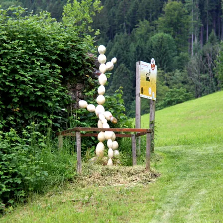 Eine menschenähnliche Figur aus gestapelten Kürbissen steht auf einem Hügel, in der Nähe eines gelben Schildes und umgeben von Grün.