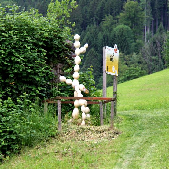 Eine menschenähnliche Figur aus gestapelten Kürbissen steht auf einem Hügel, in der Nähe eines gelben Schildes und umgeben von Grün.