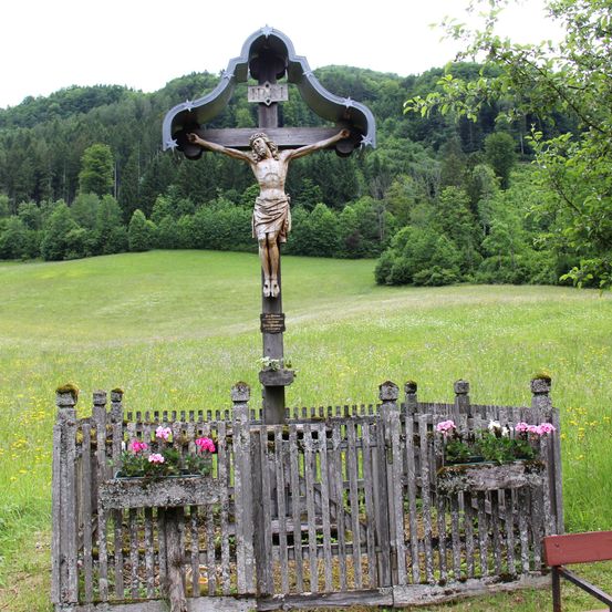 Ein hölzernes Kruzifix steht auf einem ländlichen Feld mit einem Holzzaun und blühenden Blumen. Das Kruzifix zeigt Jesus am Kreuz mit einem Stern über ihm. Im Hintergrund sind üppige grüne Bäume und ein klarer Himmel zu sehen.