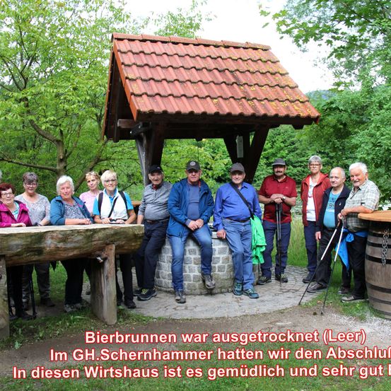 Eine Gruppe von Menschen steht vor einem hölzernen Unterstand mit einem roten Dach. Sie alle lächeln für ein Foto. Dahinter steht ein Fass und ein Baum.