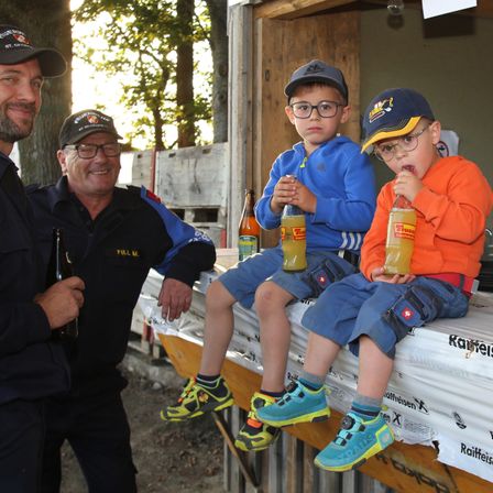 Zwei junge Jungen sitzen auf einer Bank draußen und halten Orangensaftgetränke. Sie tragen Mützen und Turnschuhe. Hinter ihnen halten zwei Männer in Uniformen Getränke. Bäume und eine Holzstruktur sind im Hintergrund.
