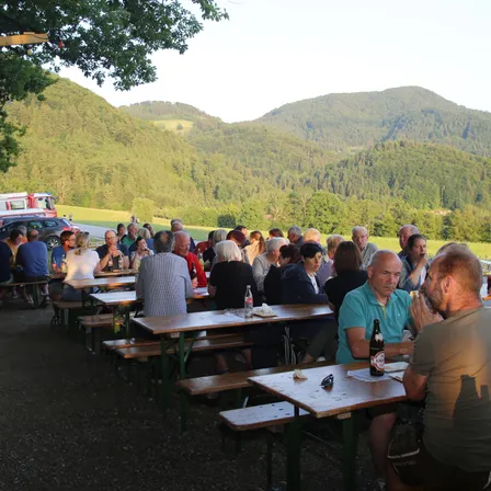 Eine Gruppe von Menschen sitzt an Holztischen in einem Außenbereich mit einer malerischen Bergsicht. Einige von ihnen klatschen, während andere trinken. Flaschen und Gläser stehen auf den Tischen.