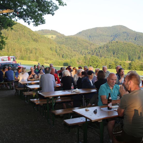 Eine Gruppe von Menschen sitzt an Holztischen in einem Außenbereich mit einer malerischen Bergsicht. Einige von ihnen klatschen, während andere trinken. Flaschen und Gläser stehen auf den Tischen.
