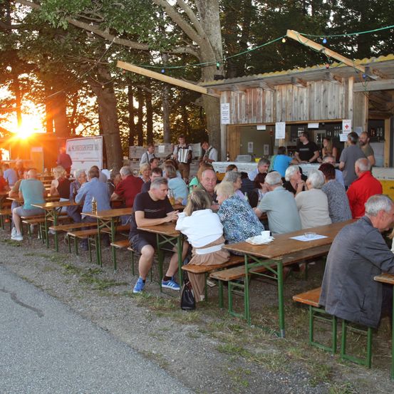 Eine Gruppe von Menschen sitzt an Bänken unter Bäumen und genießt Essen und Getränke, während die Sonne hinter ihnen untergeht. Einige Leute stehen vor einem kleinen Holzgebäude mit einer Bar.