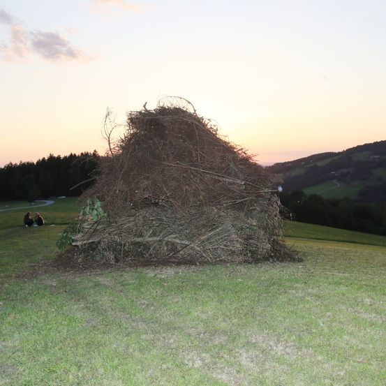 Ein Haufen Äste und Zweige auf einer grasbewachsenen Wiese bei Sonnenuntergang, mit einem Paar, das in der Nähe sitzt, und Hügeln in der Ferne.