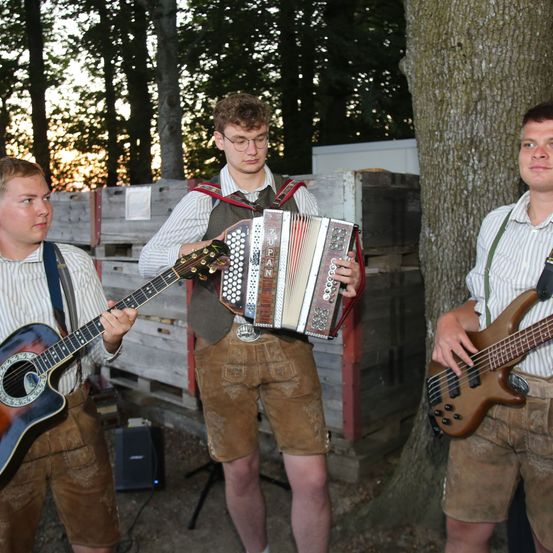 Drei Männer in traditioneller bayerischer Kleidung spielen mit ihren Instrumenten. Einer spielt Gitarre, ein anderer Akkordeon und der dritte Bassgitarre. Sie stehen vor Holzkisten und einem Baum.