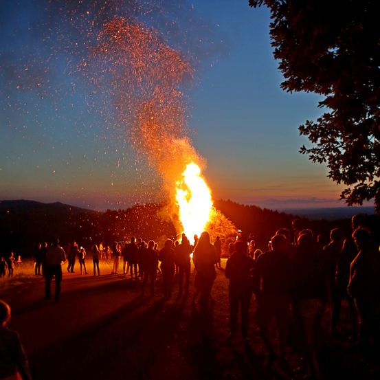Ein Lagerfeuer brennt hell in der Nacht, umgeben von Menschen. Die orangefarbenen Flammen werfen ein warmes Licht auf die Gesichter der Versammelten. Die Umgebung ist im Freien, mit Bäumen und Bergen im Hintergrund.