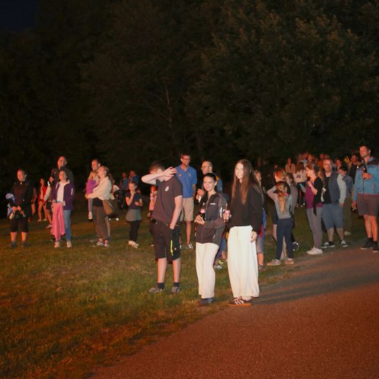 Eine Gruppe von Menschen steht auf einem Grasfeld unter dem Nachthimmel. Einige von ihnen tragen Turnschuhe und halten Flaschen. In der Ferne gibt es Bäume und eine Straße.