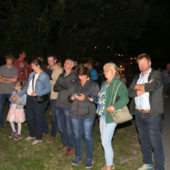 Eine Gruppe von Erwachsenen und Kindern steht auf einem Grasfeld unter einem Nachthimmel. Bäume und ein Gebäude sind im Hintergrund. Einige Leute halten Handys.