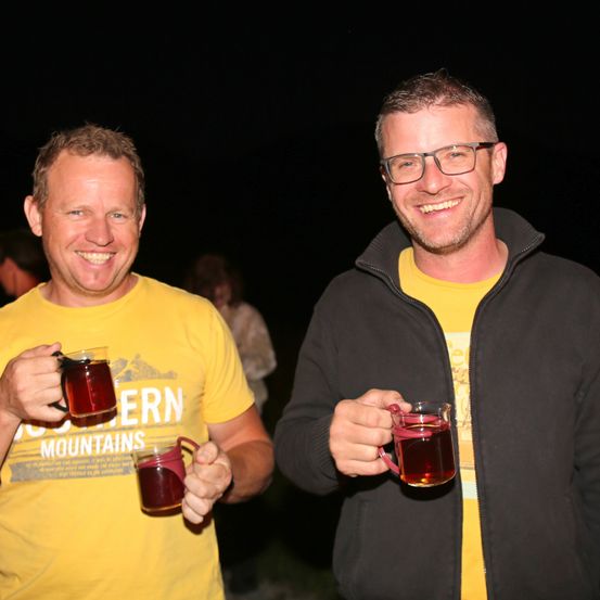 Zwei Männer, einer in einem gelben Shirt und der andere in einer schwarzen Jacke, lächeln und halten Getränke in den Händen. Der Mann im Gelben trägt eine Brille.