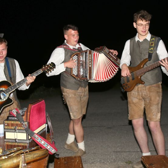 Drei Männer in traditioneller Kleidung spielen nachts draußen Instrumente. Einer spielt Akkordeon, ein anderer Gitarre und der dritte E-Bass. Im Vordergrund befinden sich Musikinstrumente und ein Tisch.