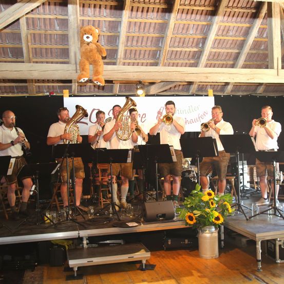 Eine Gruppe von Musikern spielt auf einer Bühne mit Musikinstrumenten, darunter Trompeten und Tubas. Ein Teddybär hängt von der Decke. Sonnenblumen stehen in einem Glas auf der Bühne.