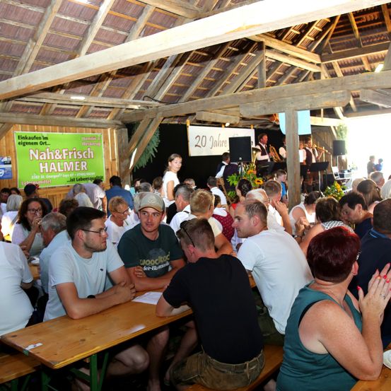 In einem großen Saal sitzen viele Menschen an Tischen und Bänken und lauschen einer Band auf der Bühne. Ein Banner im Hintergrund trägt die Aufschrift 'Nah&Frisch HALMER'.