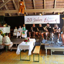 Eine Gruppe von Musikern spielt Instrumente auf einer Bühne, mit einem Banner, das '20 Jahre Brundler Blechhauvon' steht. Kinder sitzen auf der Bühne, und ein Priester steht vor ihnen.