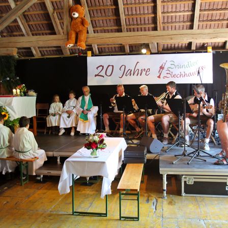 Eine Gruppe von Musikern spielt Instrumente auf einer Bühne, mit einem Banner, das '20 Jahre Brundler Blechhauvon' steht. Kinder sitzen auf der Bühne, und ein Priester steht vor ihnen.
